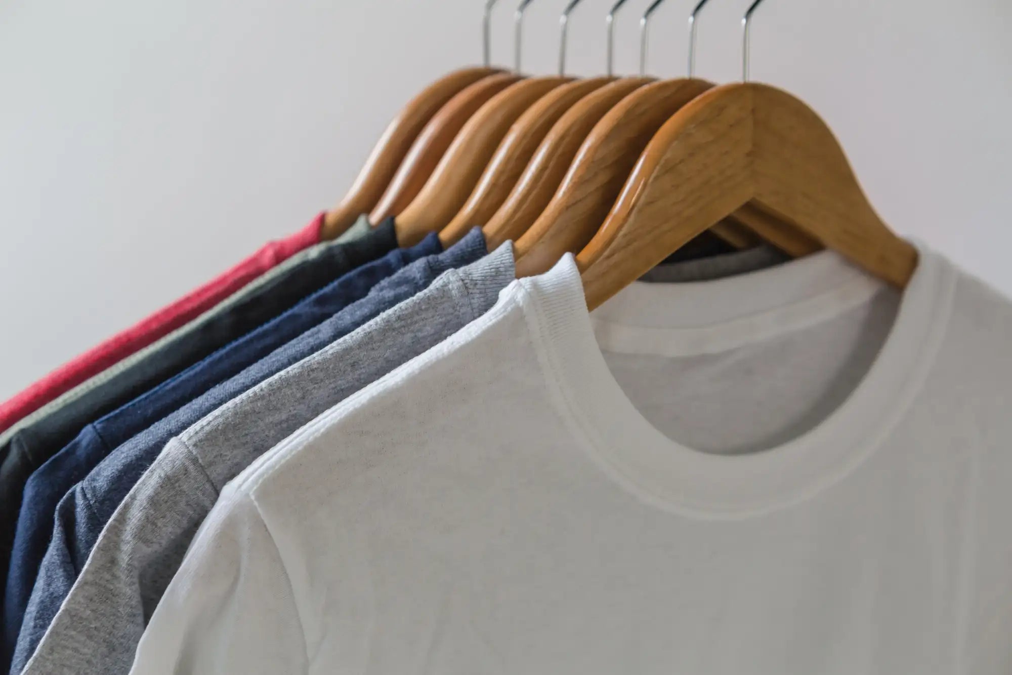 A white cotton crew-neck t-shirt hanging on a wooden hanger, with other colored shirts visible behind it.